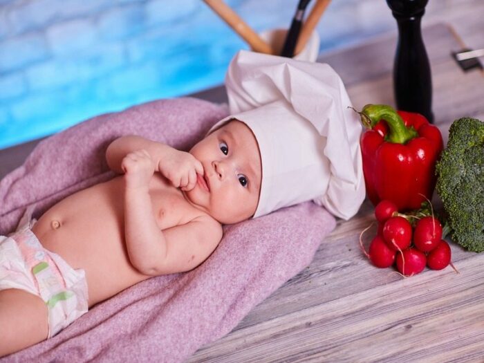 Bébé allongé sur une serviette avec une toque de cuisinier sur la tête. A côté de lui il y a un poivron rouge, un brocoli et des radiset